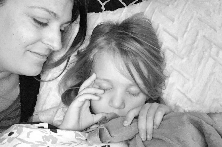 a mom laying next to her daughter who is asleep and sucking her thumb and holding her lovey