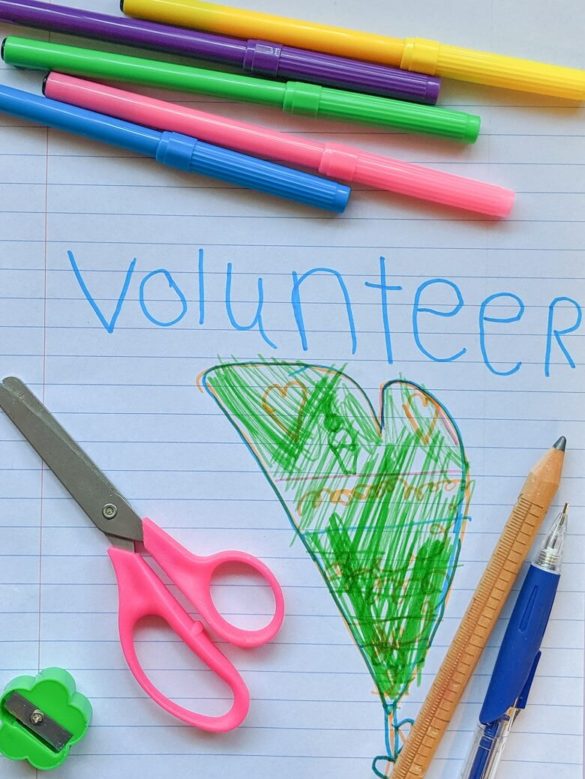 Notebook paper with the word "volunteer" written in children's writing and a picture of a heart drawn with markers surrounded by school supplies like pencils, markers, scissors and a pencil sharpener