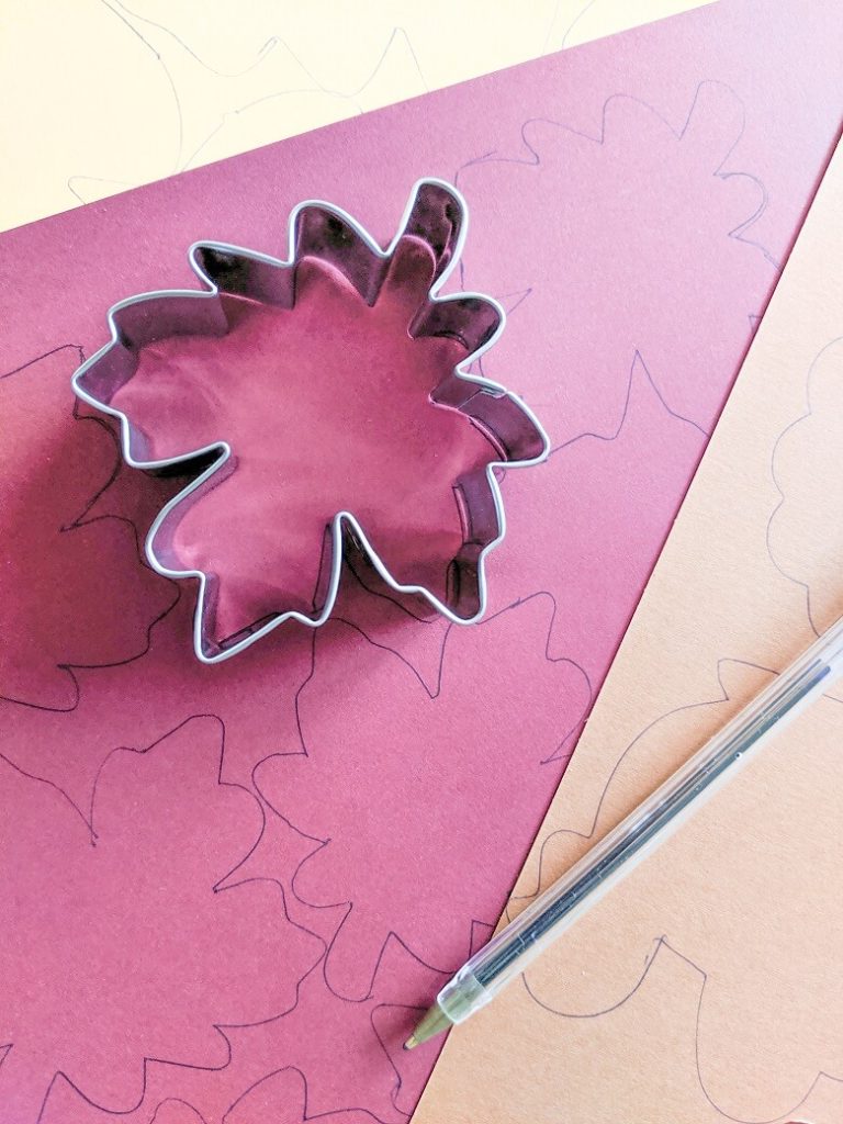 Materials for a thankfulness craft that include a maple leave cookie cutter on maroon paper with a pen. The paper has traced maple leaves on it and there is a yellow piece of paper with traced leaves and a brown piece of paper with traced turkey shapes on it.