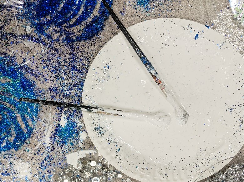 Silver and blue glitter on plates and spilled around a table alongside another plate with glue and two paint brushes