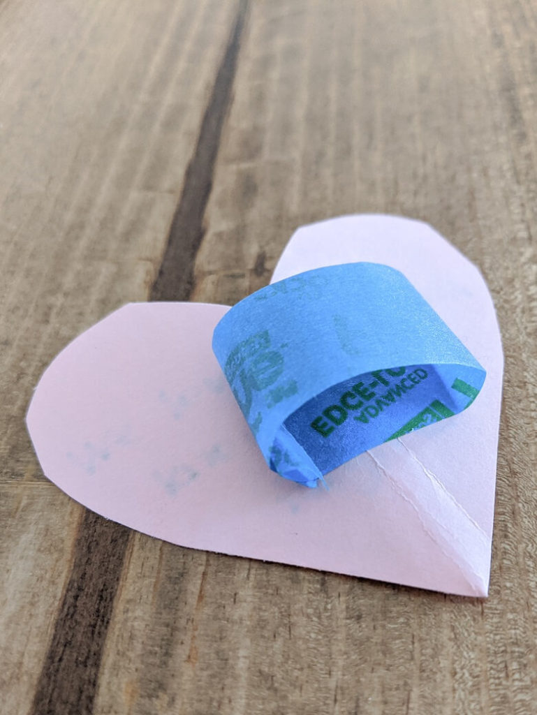 A heart cut out of construction paper with a piece of painter's tape rolled up on the back and a love message on the front which is facing down.