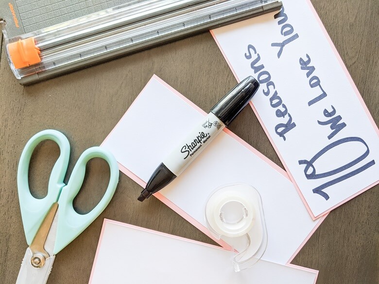 Some materials needed to make this Valentine's Day craft: Scissors, tape, permanent marker, paper cutter, and a sign that says 10 Reasons We Love You