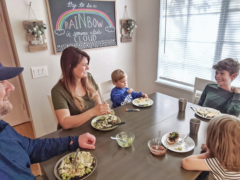 A smiling family of 5 with a mom and dad, young boy and girl, and a toddler around a table in a dining room eating a plant-based dinner.