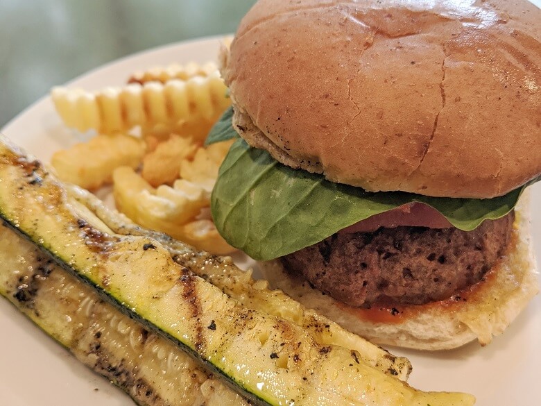 A vegan family meal consisting of a Beyond Burger with tomato, spinach, avocado on a bun next to zucchini and french fries