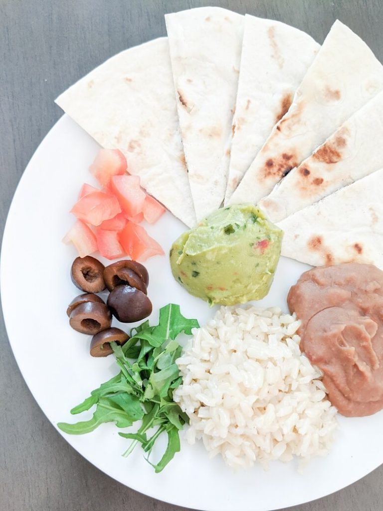 Showing a kid-friendly vegan dinner with a kid's plate of a vegan taco dinner consisting of a tortilla cut into triangles, guacamole, beans, rice, lettuce, olives and tomatoes
