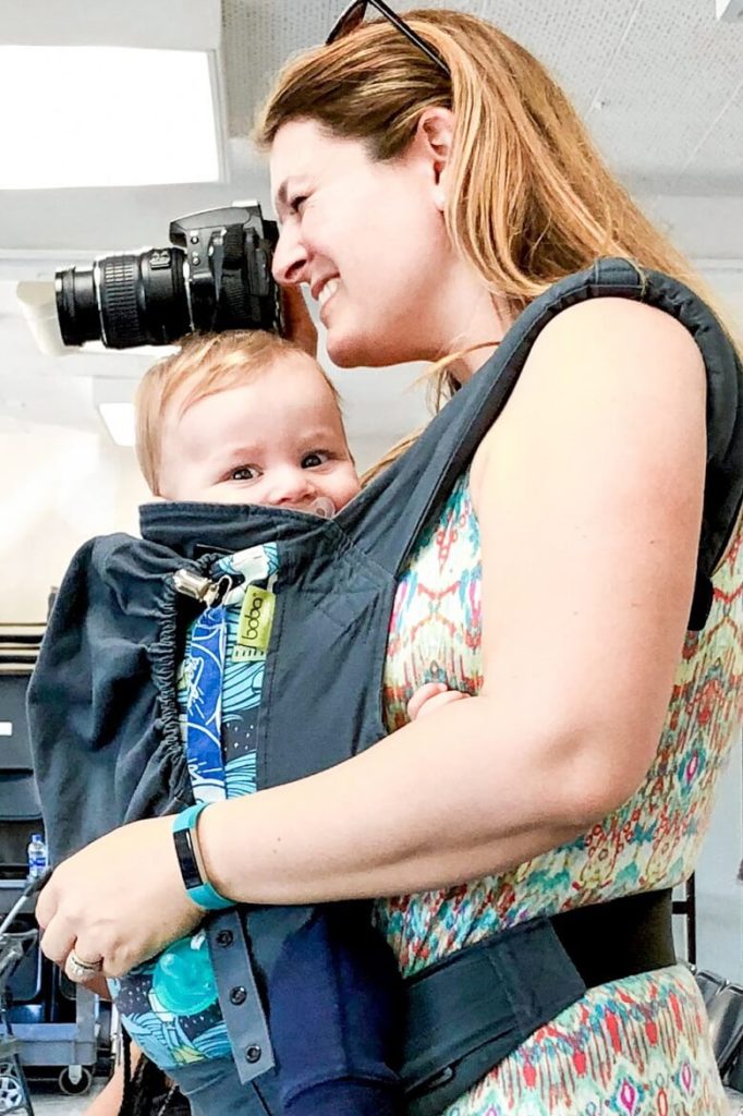 A school volunteer taking pictures with her camera while wearing a young toddler in a baby carrier.