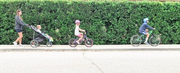 A mom showing one of the fun ways to workout at home as a family by walking in the neighborhood with her three kids. She pushes one in a stroller and the other two are riding bikes.