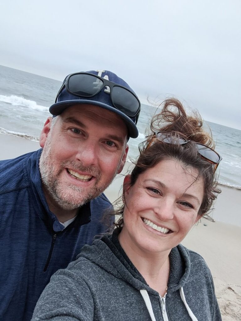 A couple in a loving relationship smiling at the camera with the beach in the background