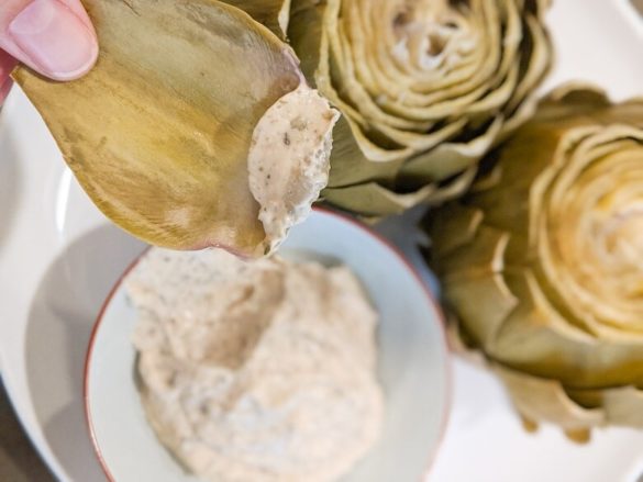 A person holding an artichoke leaf with a vegan artichoke dipping sauce on it. Two artichokes and a bowl of dip are in the background.