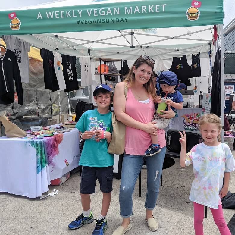 A vegan family consisting of a mom holding a toddler and two other young children all standing in front of a vegan market