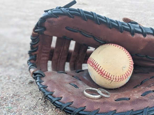 A glove on a ball field with a baseball in the center and two wedding rings in front of it.