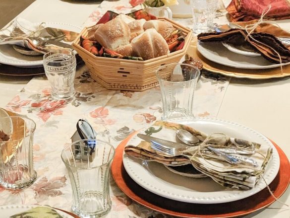 A dinner table set for Thanksgiving with place settings, silverware, cups and rolls in a basket and autumn decor.