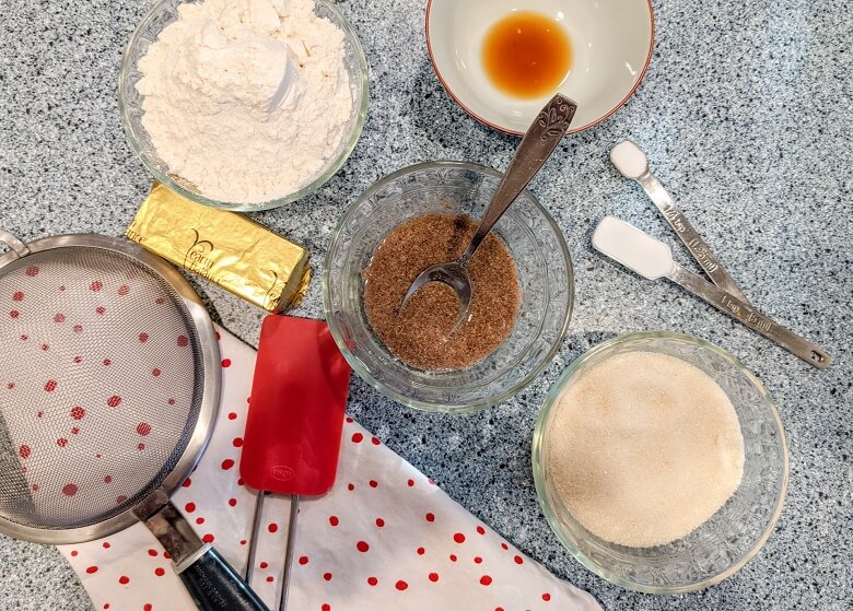 Vegan sugar cookie ingredients on a table, which include flour, Earth Balance butter, vanilla extract, flax egg, sugar, baking soda, salt, and a spatula and sifter are also shown.