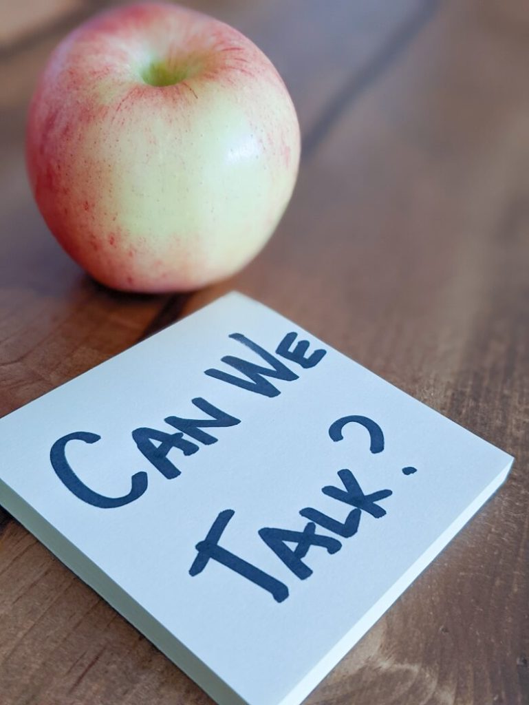 an apple on a table next to a notepad that reads "Can we talk?"