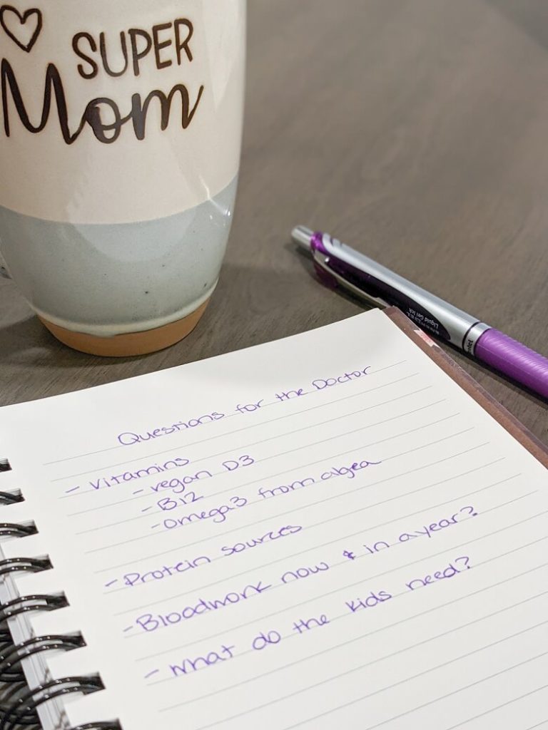 A notepad of questions to ask a doctor for a vegan beginner that reads: Vitamins, vegan D3, B12, Omega 3 from algea; Protein sources; Bloodwork now and in a year?; what do the kids need? There is also a pen and a mug that reads "super mom" in the background