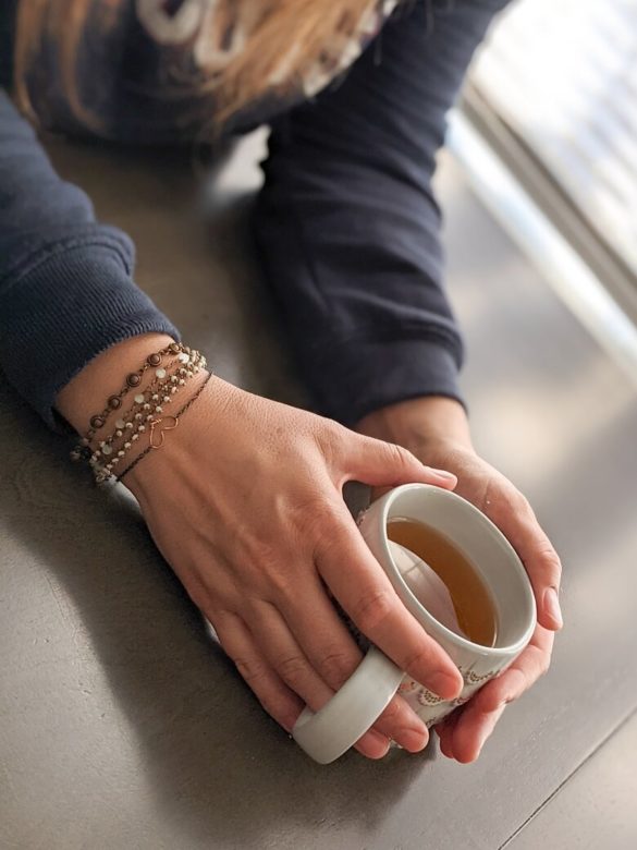 A woman's hands holding a cup of tea on a table. One of her hands has a bracelet with a heart on it to represent compassion as she is supposed to be talking to someone about going vegan.
