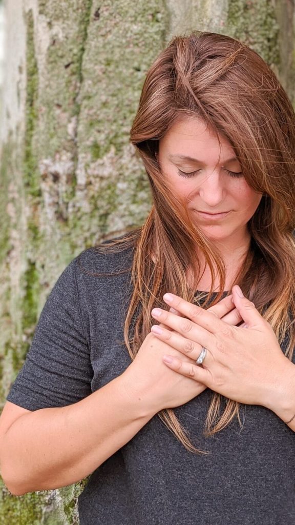 A photo of a woman who looks calm and meditative with her eyes closed and head bent with her hands over her heart.
