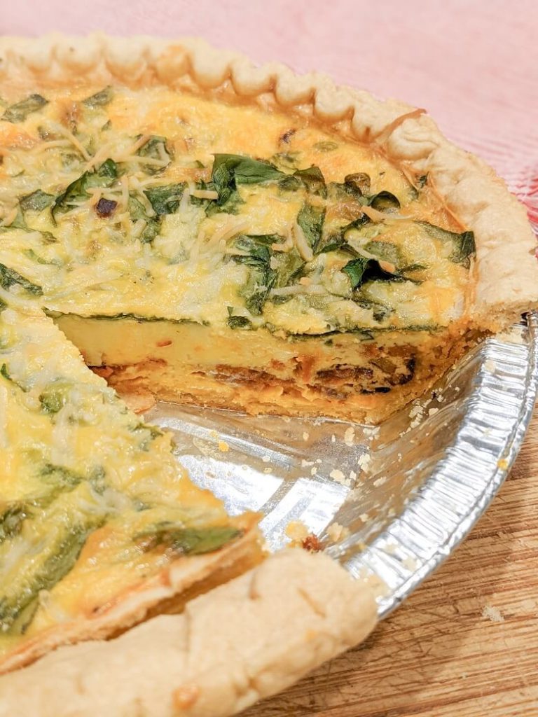 A cooked vegan quiche with a slice cut out of it showing the layers inside of spinach, vegan bacon and melted cheese and cooked JUST Egg.