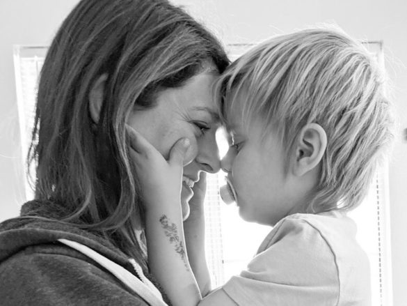 A photo of the profile of a mom and her young son touching noses. The child's hands are on the mom's cheeks. They are in front of a window.