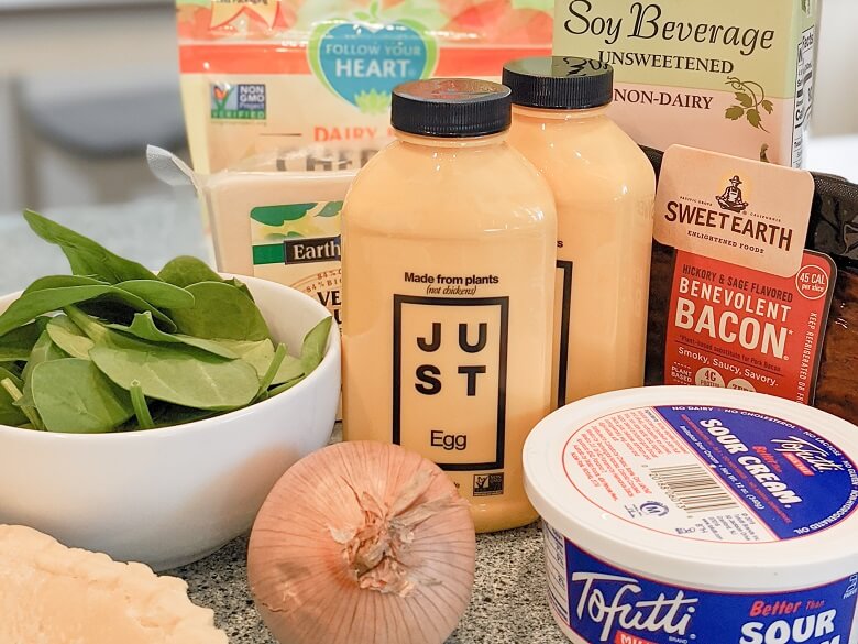 Ingredients for a vegan quiche recipe which include: pie shell, onion, vegan sour cream, spinach, JUST Egg, Sweet Earth Benevolent Bacon, soy milk, and vegan cheeses like Follow Your Heart vegan cheddar