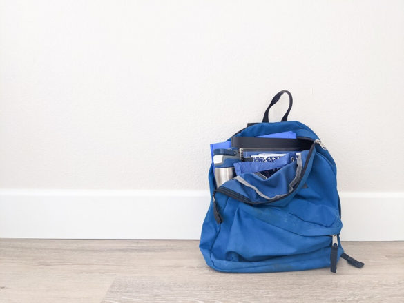 A photo of a half unzipped backpack against a wall with notebooks, a water bottle and pencil pouch slightly popping out.