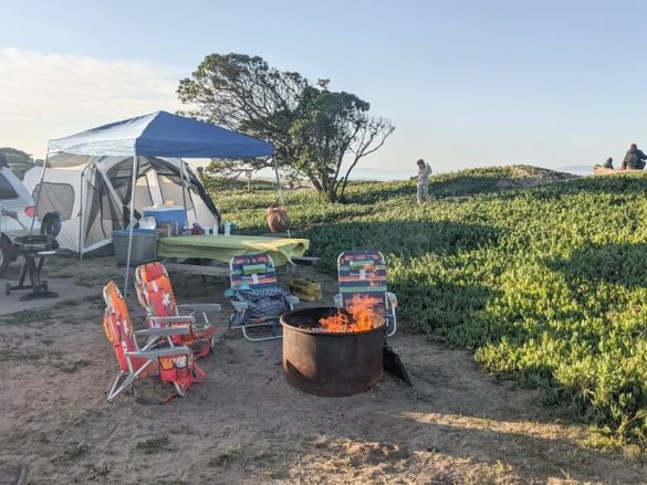 A beach camp ground with a fire in the fire pit, chairs surrounding it, a picnic table with an easy up over it, a tent, a portable stand up barbecue and a child running in the background