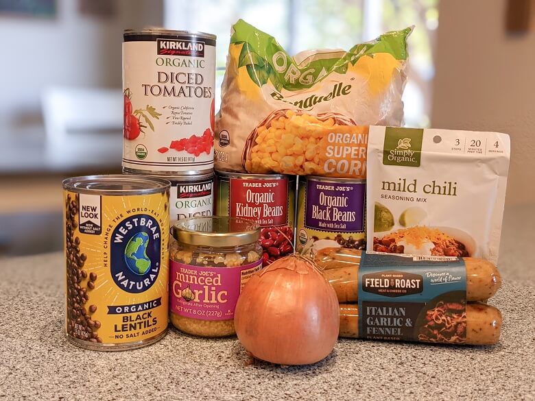 Ingredients for a vegan chili recipe on a kitchen counter that includes a can of lentils, two cans of diced tomatoes, a can of red kidney beans, a can of black beans, a jar of minced garlic, an onion, a bag of frozen corn, Field Roast vegan sausages and a packet of Simply Organic's Mild Chili Seasoning Mix
