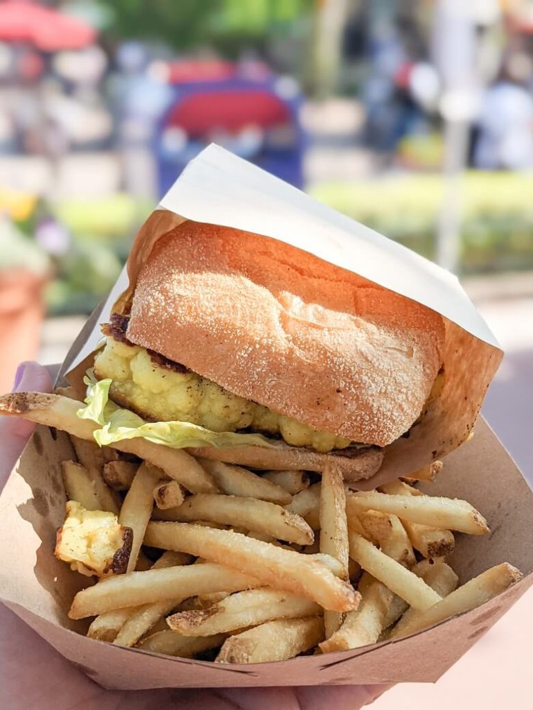 The cauliflower sandwich with fries in a paper tray with Fantasy Land in the background.
