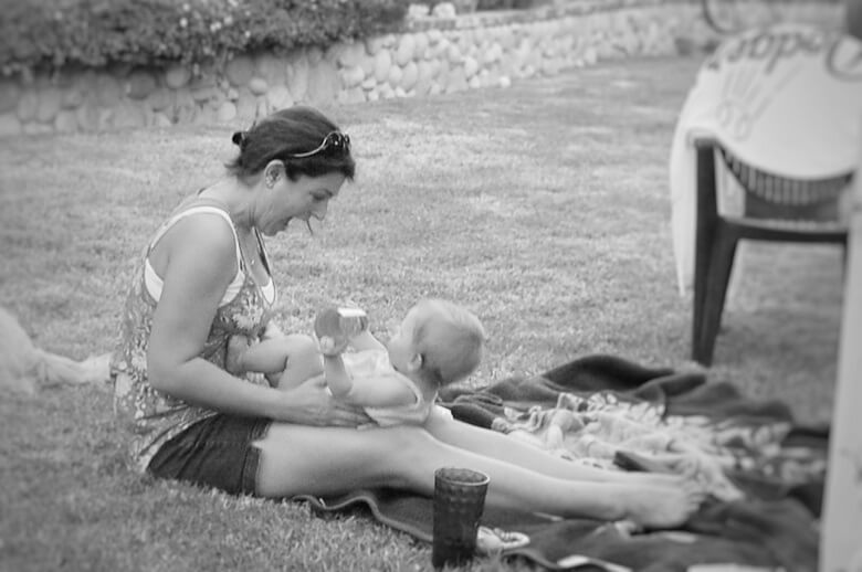 A SAHM holding her baby in her lap and laughing while they play. They are sitting on a blanket on the grass.