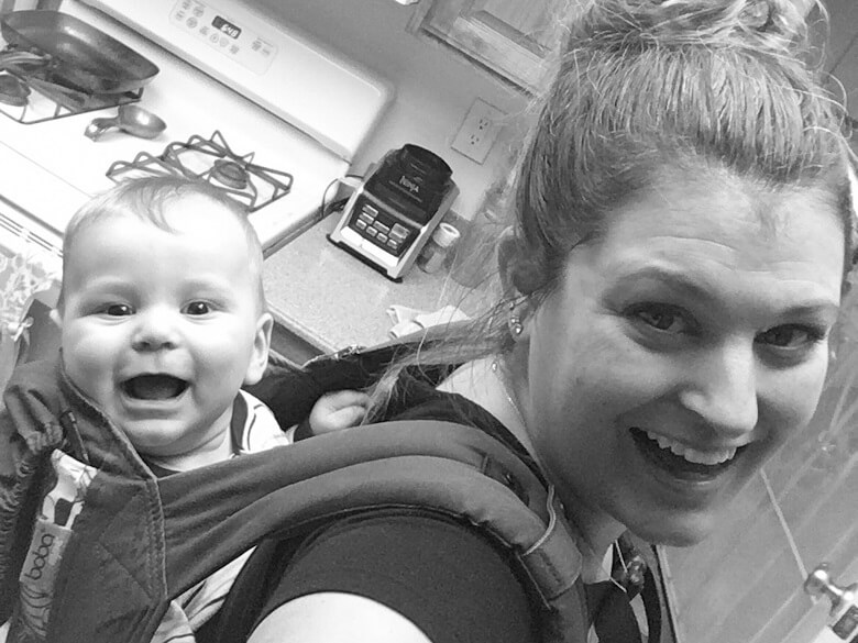 A stay-at-home mom in her kitchen wearing her baby on her back in a carrier.