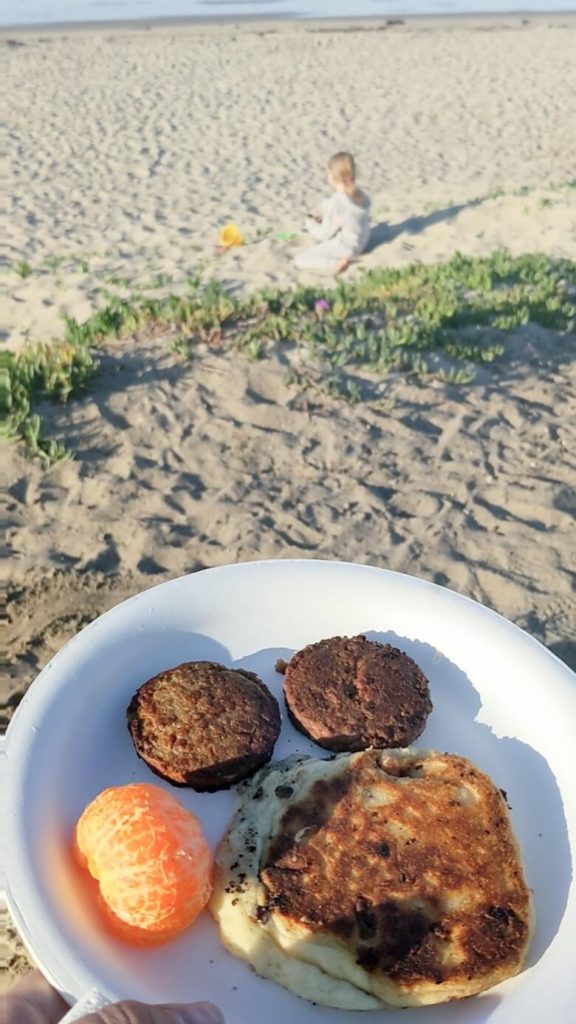 Vegan breakfast food at a beach campground that includes a Mandarin orange, two Beyond Breakfast Sausages and a chocolate chip pancake on a paper plate with a young girl playing on the sand and the ocean in the distance.
