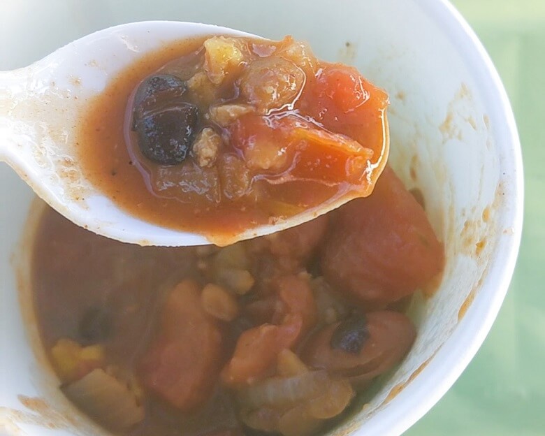 A plastic spoon holding a scoop of vegan chili over a paper cup with more chili on a camp group table.