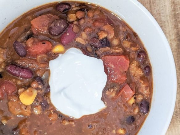 Bowl filled with vegan chili that includes diced tomatoes, black beans, red kidney beans, corn, lentils, and a dollop of vegan sour cream in the center.