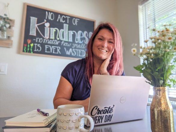 An image of a woman who is blogging is sitting at her laptop, smiling at the camera with flowers in the foreground and a chalkboard in the background that says "No act of kindness no matter how small is ever wasted"