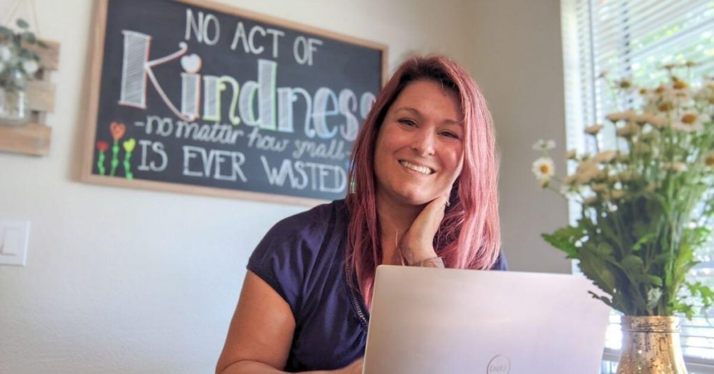 An image of a woman who is blogging is sitting at her laptop, smiling at the camera with flowers in the foreground and a chalkboard in the background that says "No act of kindness no matter how small is ever wasted"