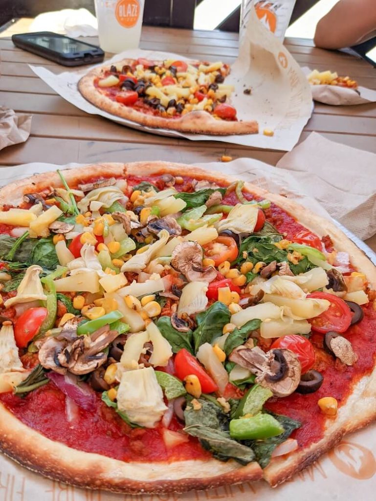 Vegan pizza from Blaze Pizza that has two pizzas with lots of vegetables on top and no cheese. Both pizzas are on a table with drinks in the background.