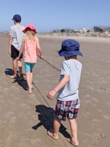 Three children mindfully walking in a line on the sand on a beach with the view from their backs. An older boy in a hat is at the front, followed by a girl who is a little younger and a preschool age boy at the back. They all are holding a long strand of seaweed.