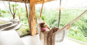 A woman laying in a hammock that's positioned between two posts, under a wooden patio. She's wrapped in several thin blankets and looking out over a peaceful, green rainforest.