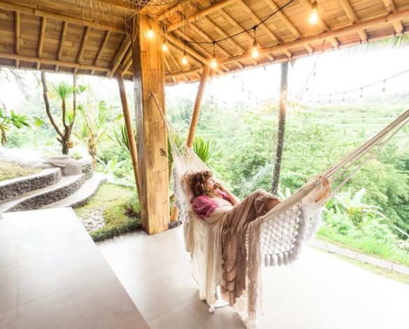 A woman laying in a hammock that's positioned between two posts, under a wooden patio. She's wrapped in several thin blankets and looking out over a peaceful, green rainforest.
