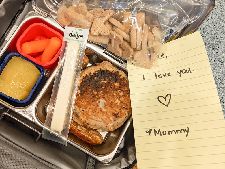 A vegan lunch for kids that includes a bag of alphabet cookies, a Daiya cheese stick, pancakes and plant-based sausage, two carrot sticks and gold kiwi. A not from mom that says "I love you. Heart, Mom" is also included.