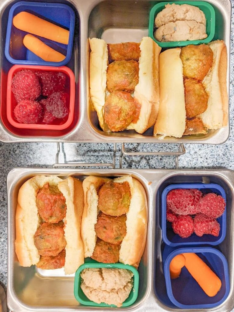 Two stainless steel vegan lunch boxes with the same food in each compartment: two cookies, two carrot sticks, a handful of raspberries, and two small meatless meatball sandwiches