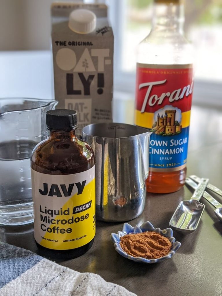 The ingredients needed to make the copy cat iced brown sugar oatmilk shaken espresso at Starbucks. From left to right, a glass of water, a bottle of Javy Coffee concentrate, a box of Oatly oat milk, a small steel pitcher for milk, a bottle of Torani brown sugar cinnamon syrup, a small dish with ground cinnamon and three teaspoons.