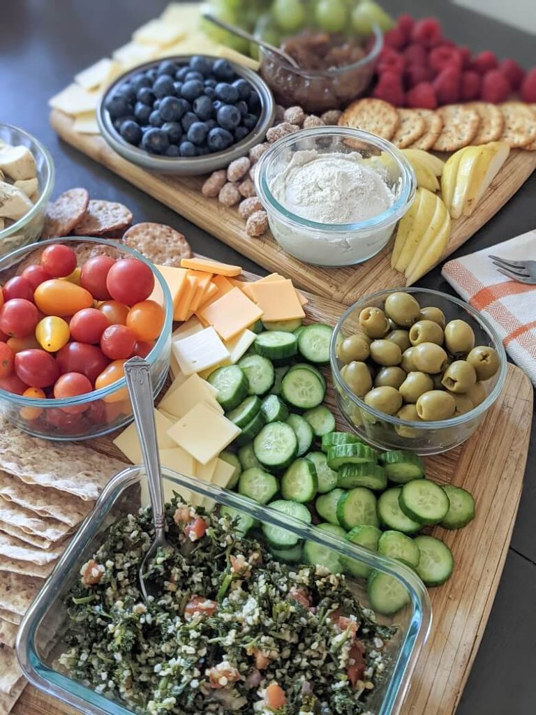 Two wooden cutting boards on a table. The one further away has vegan dip, jam, almonds, pears, crackers, raspberries, blueberries, grapes and vegan cheese. The one closer has tabouli, Persian cucumbers, green olives, vegan cheeses, lavash, grape tomatoes, crackers and marinated artichoke hearts.