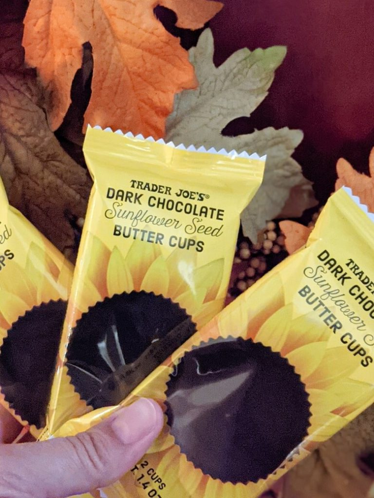 Fall foliage in the background with a hand holding three packages of Trader Joe's Dark Chocolate Sunflower Seed Butter Cups