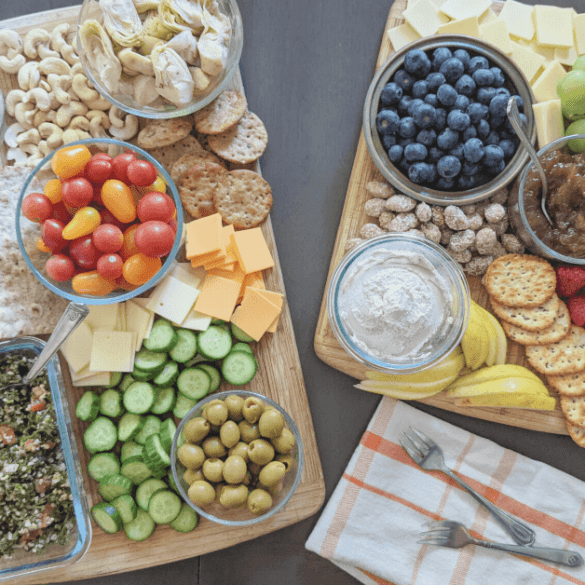 Two wooden cutting boards on a table. One has vegan dip, jam, almonds, pears, crackers, raspberries, blueberries, grapes and vegan cheese. The other one has tabouli, Persian cucumbers, green olives, vegan cheeses, lavash, grape tomatoes, crackers, marinated artichoke hearts, hummus and cashews.