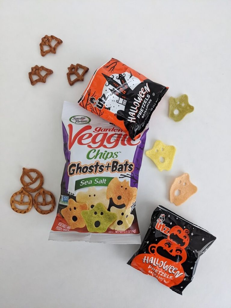 A bag of Garden Veggie Chips Ghosts and Bats with a few out of the bag and two bags of Halloween-shaped pretzels with a few bat and pumpkin shaped ones out of the bag.