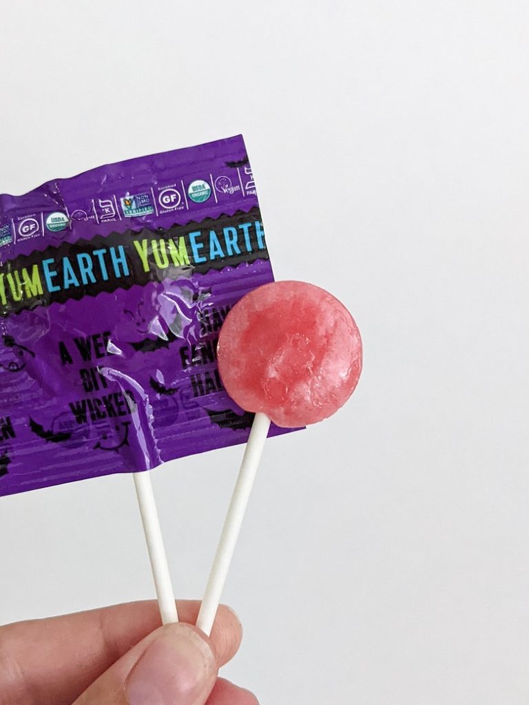 A hand holding up two YumEarth lollipops. One in a wrapper and one opened.