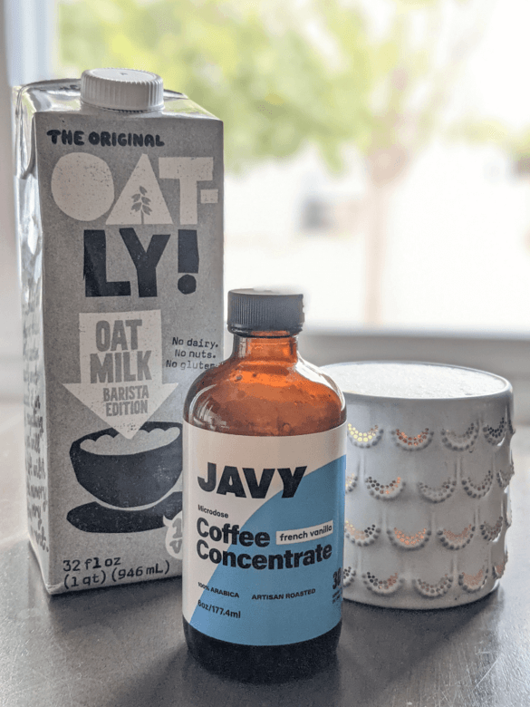 A box of Oatly barista edition oat milk, a jar of Javy Coffee Concentrate and a mug of coffee filled with a latte on a table.