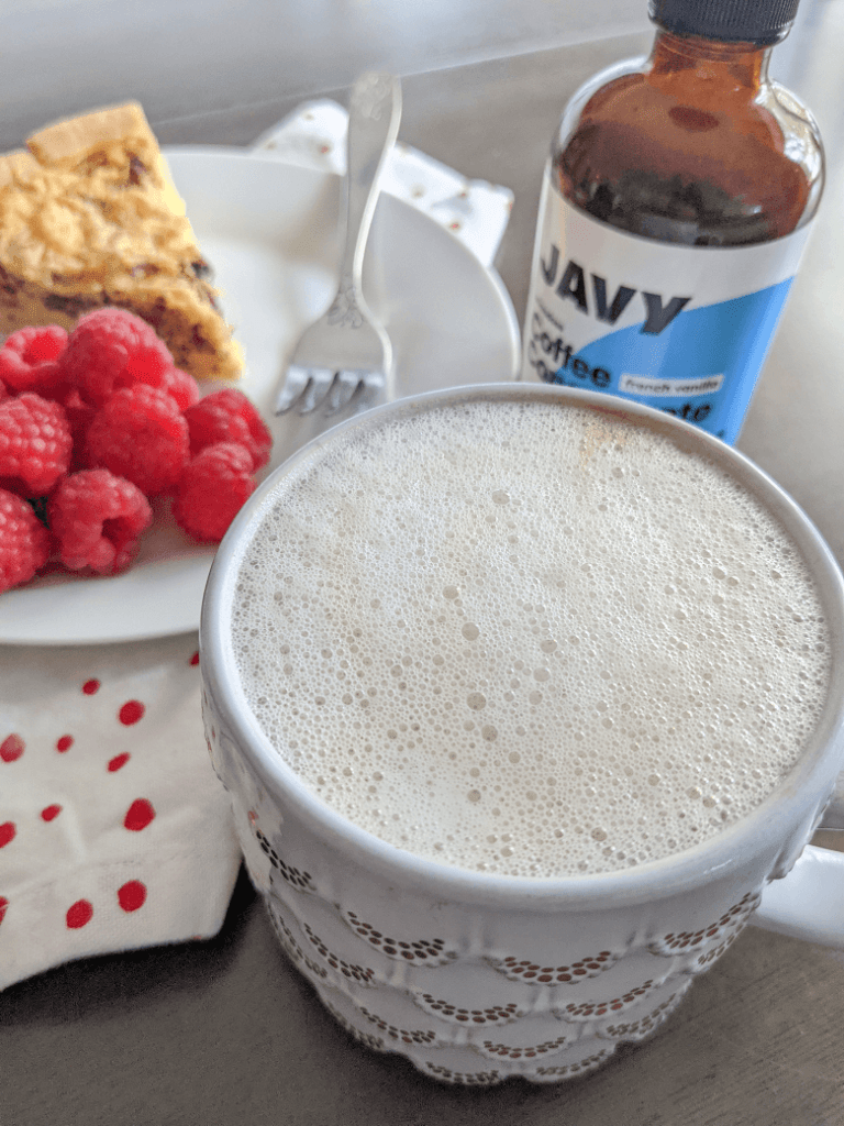 A mug with a latte in it next to a bottle of Javy Coffee concentrate, a napkin, a plate with a fork and vegan quiche and raspberries.
