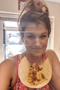 A woman smiling at the camera holding a tortilla filled with plant-based food like tofu, spinach, potatoes and salsa.